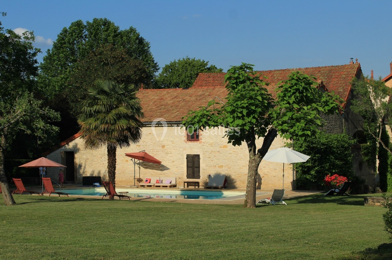 Maison en pierre avec piscine, transats et parasols dans un jardin arboré par une journée ensoleillée.