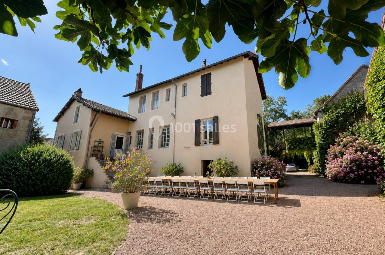 Grande maison beige avec volets en bois, entourée de végétation, et longue table extérieure dressée sur une allée…
