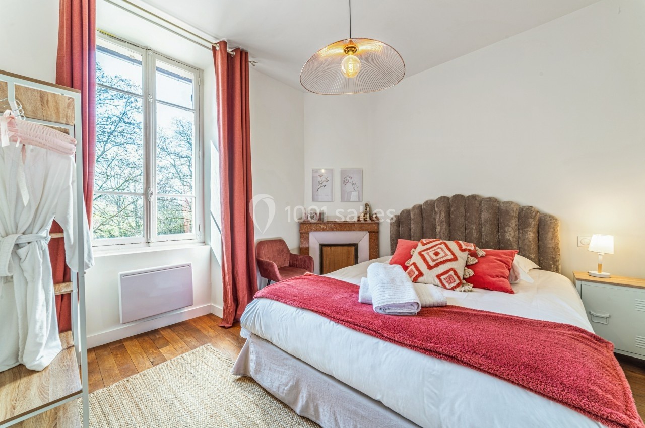 Chambre lumineuse avec lit double, linge de maison rouge et blanc, grande fenêtre et décoration chaleureuse.