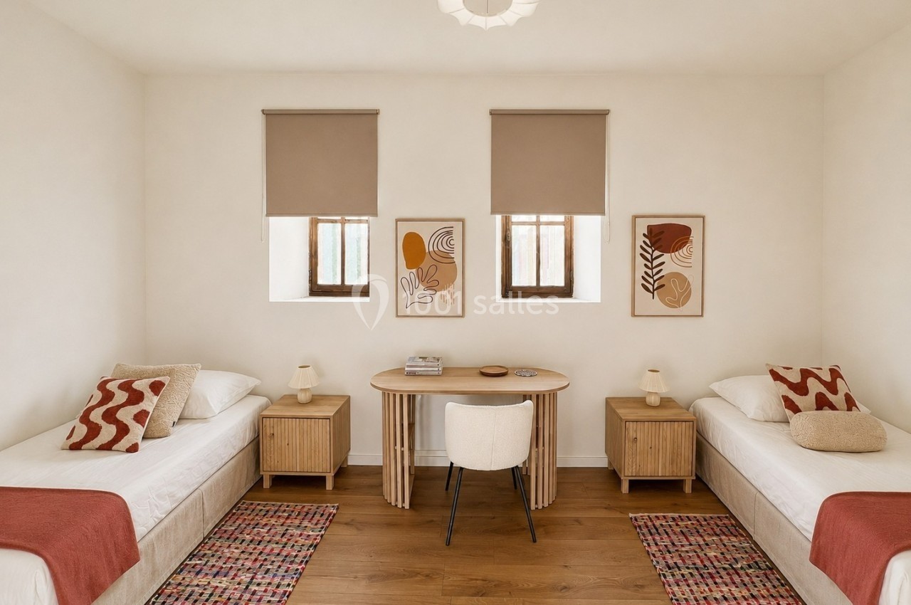 Deux lits simples avec linge rouge, table centrale en bois, chaises et décorations murales dans une chambre lumineuse.