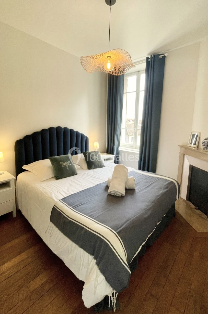 Chambre lumineuse avec lit double, draps pliés, parquet en bois, cheminée et grande fenêtre avec rideaux sombres.