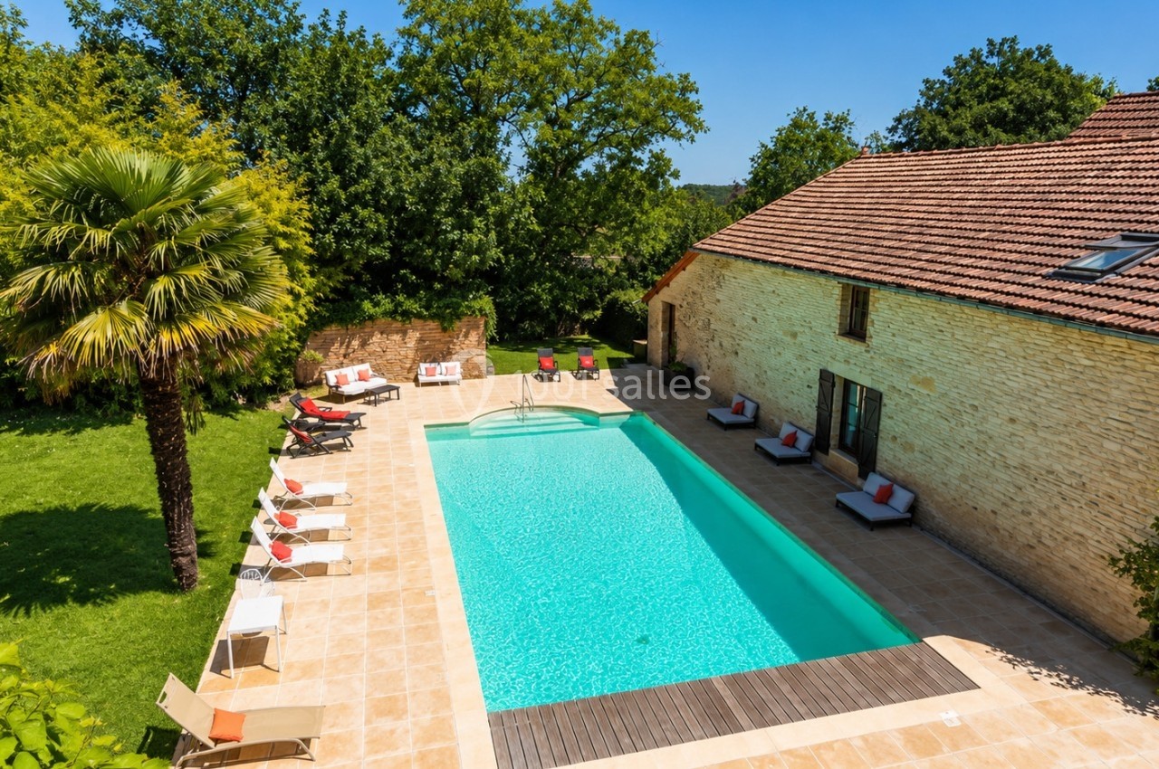 Vue d'une maison en pierre avec une piscine entourée de transats, située dans un jardin arboré.