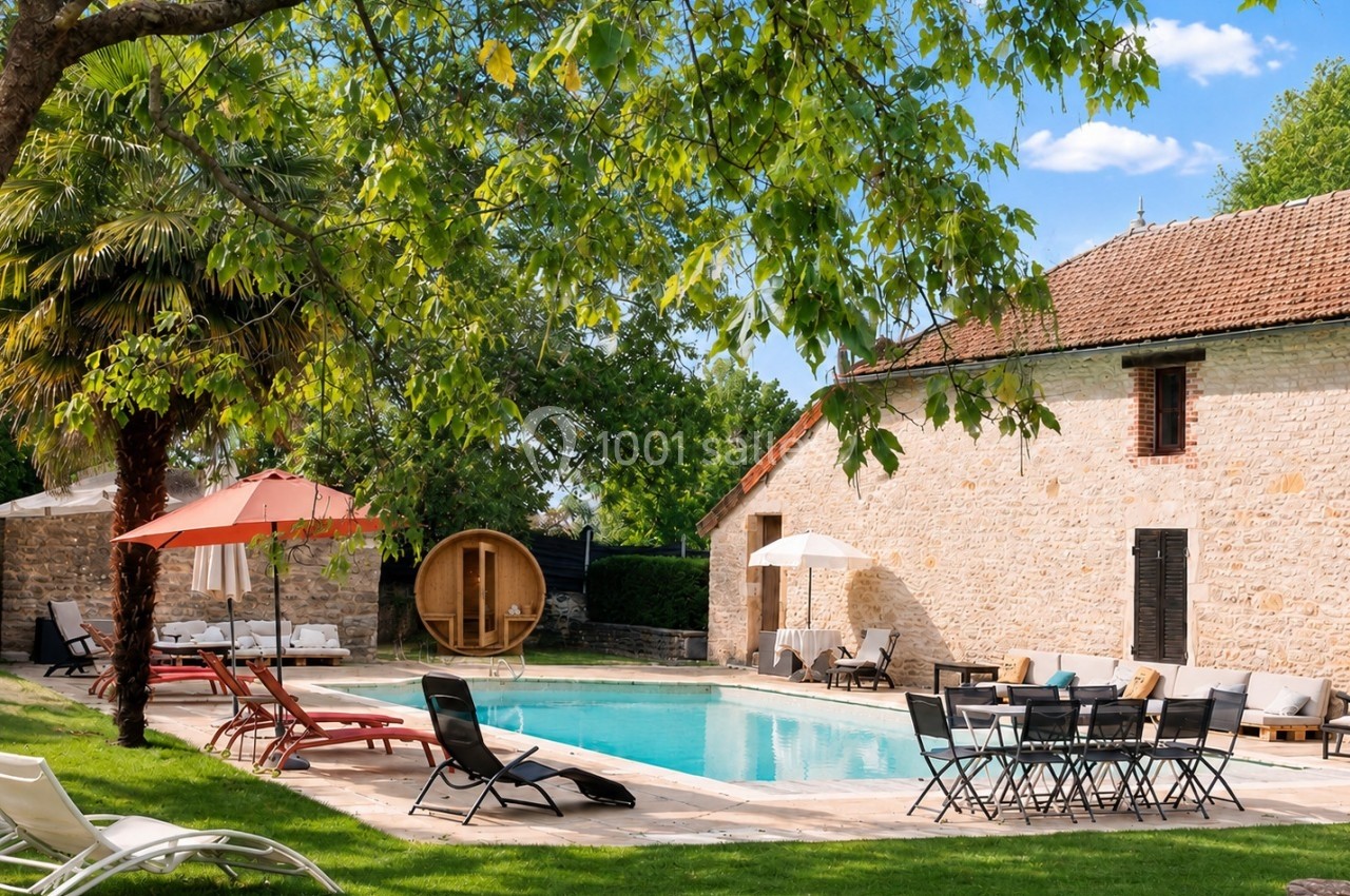 Piscine extérieure entourée de chaises longues, parasols, sauna en bois et bâtiment en pierre sous un ciel dégagé.