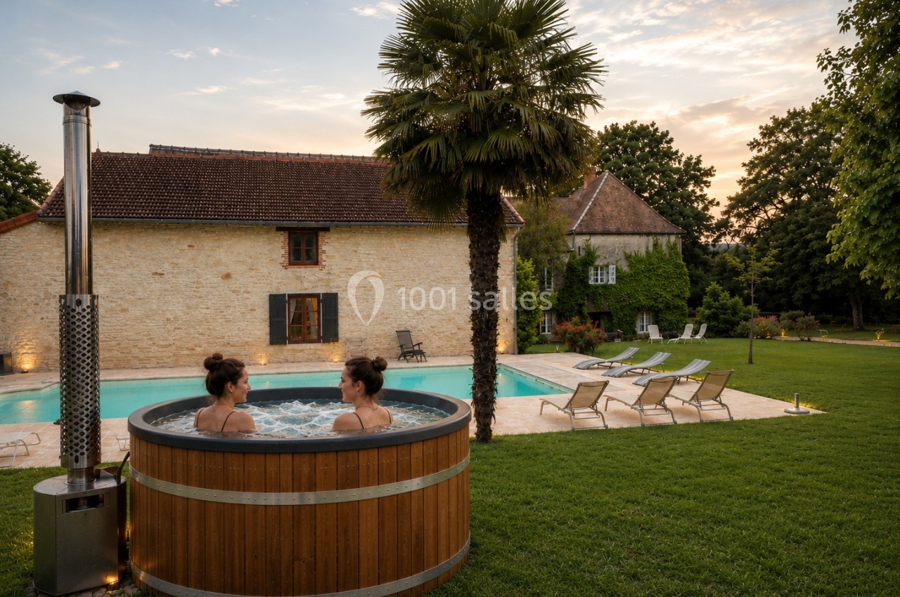 Deux personnes dans un bain nordique en bois près d'une piscine, entourées de verdure et d'une maison en pierre au coucher…