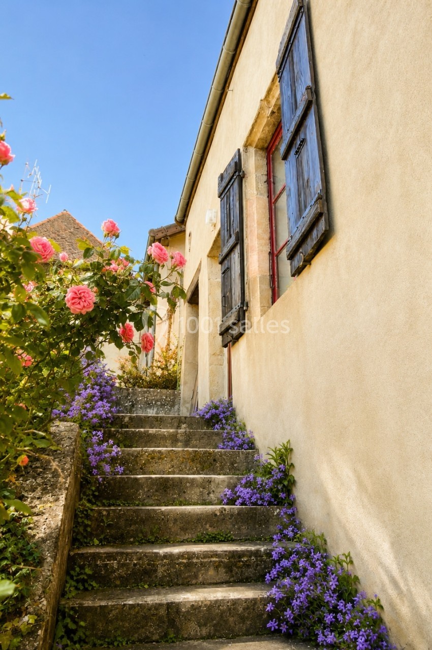 Escalier en pierre bordé de fleurs violettes et roses menant à une maison aux volets en bois sous un ciel bleu.
