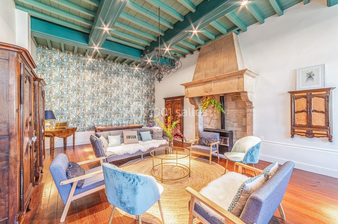 Salon lumineux avec plafond en poutres vertes, cheminée en pierre, mobilier moderne et décoration florale.