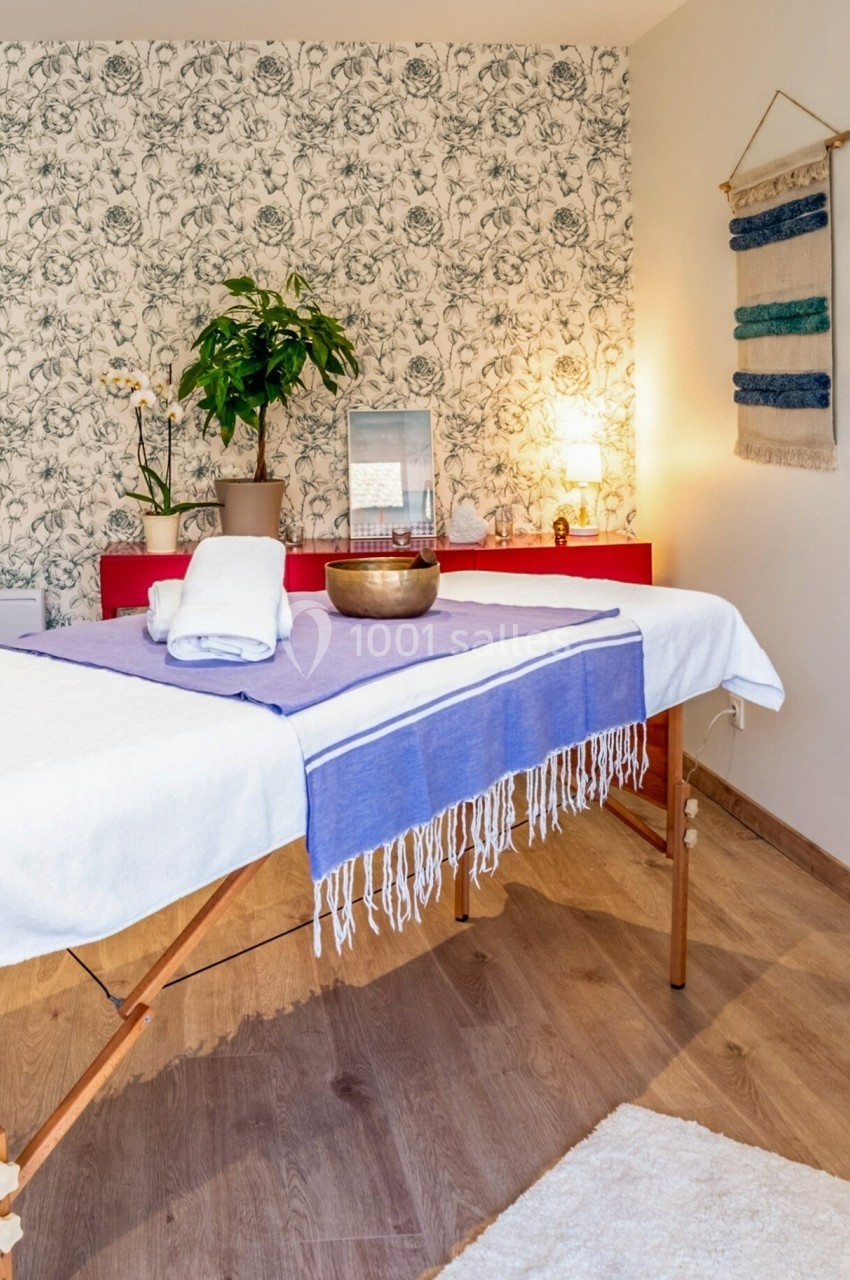 Table de massage avec serviettes pliées, bol tibétain et décoration murale dans une pièce lumineuse et apaisante.