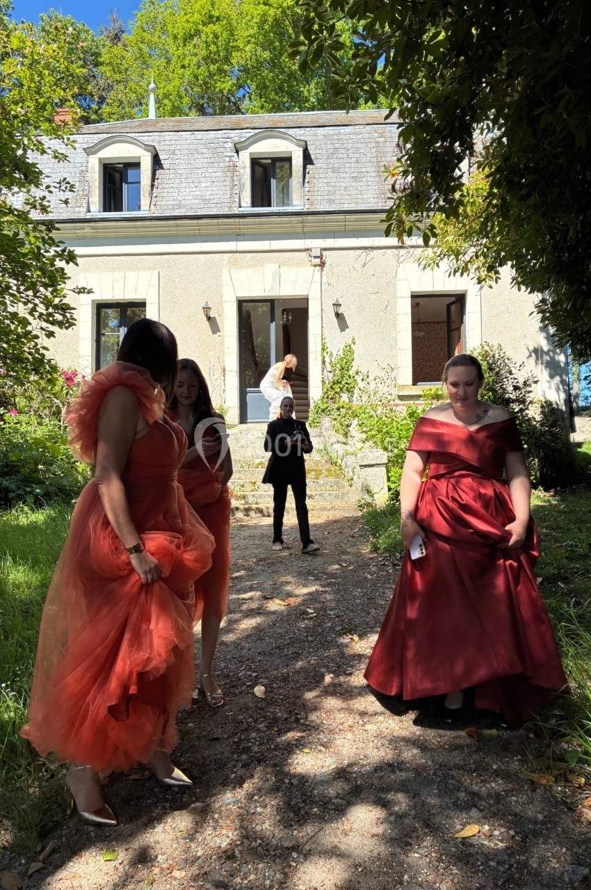 Quatre personnes en robes élégantes discutent devant une maison en pierre entourée de verdure.