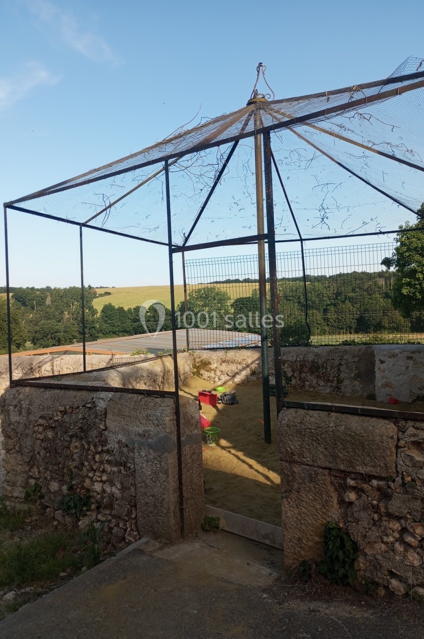 Enclos grillagé ouvert, situé dans une cour en pierre, avec vue sur un paysage rural et des arbres.