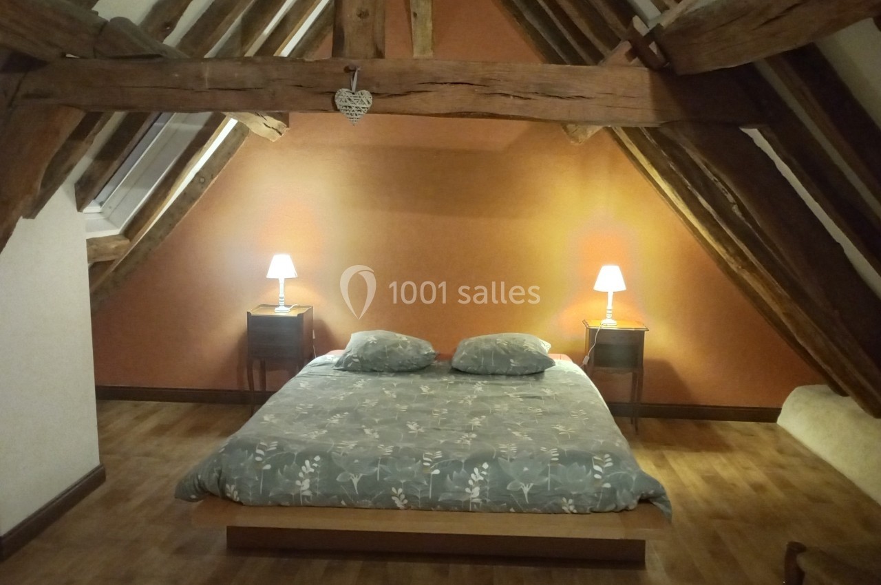 Chambre sous combles avec poutres apparentes, lit double, deux lampes de chevet allumées et parquet au sol.
