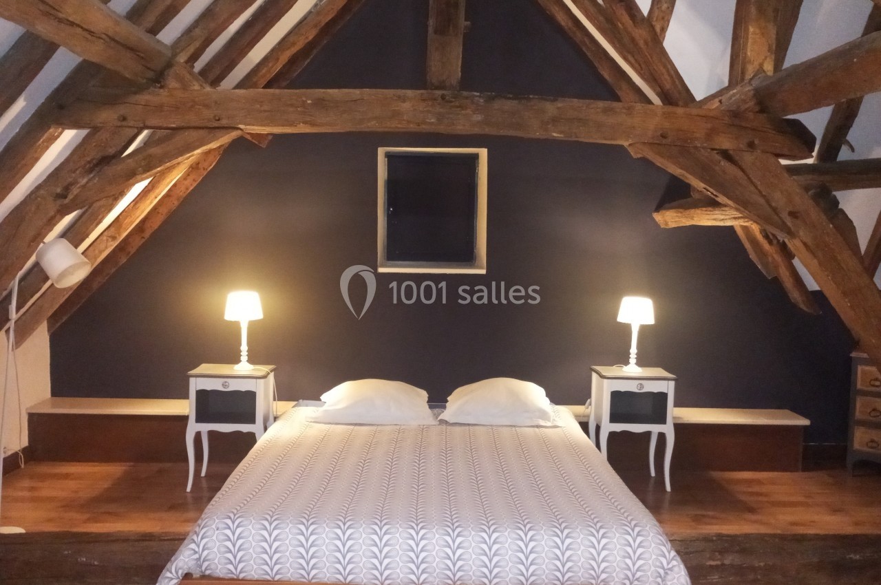 Chambre sous combles avec poutres apparentes, lit double, deux lampes de chevet et mur peint en bleu foncé.
