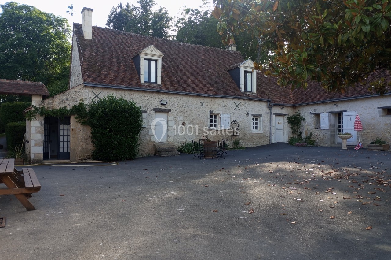 Cour pavée avec maison en pierre, toit en tuiles, mobilier de jardin et arbres en arrière-plan.