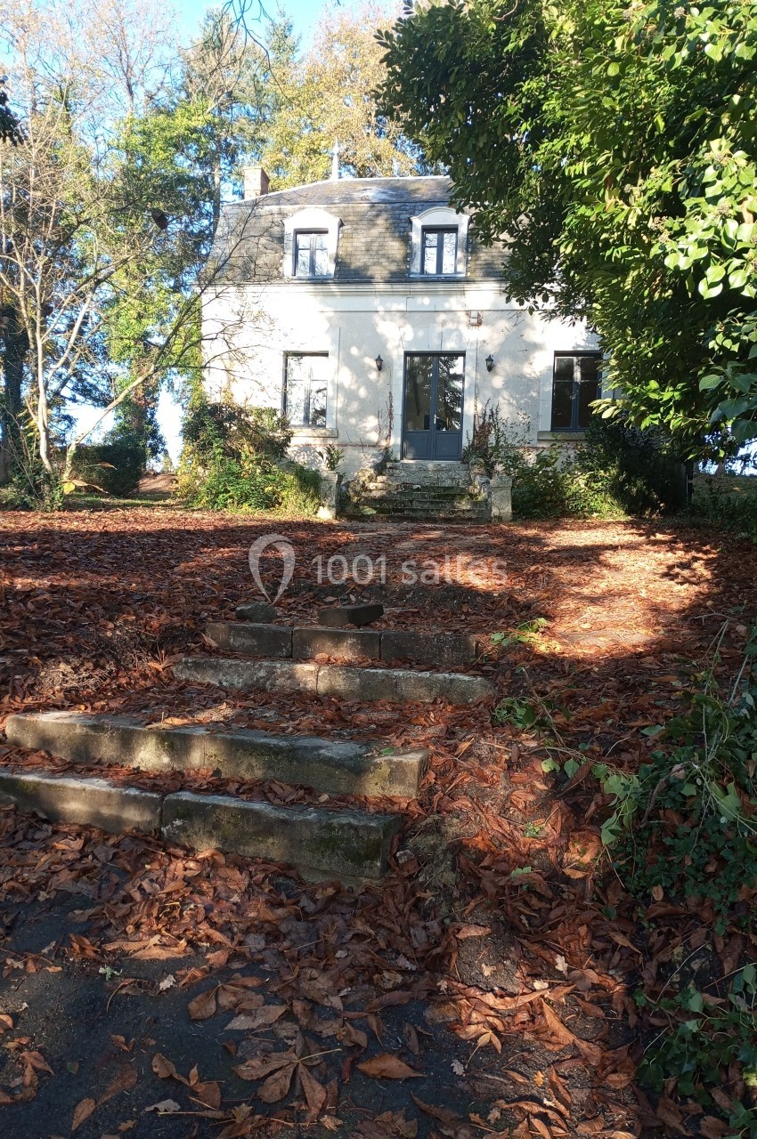 Maison blanche entourée d'arbres, avec des marches en pierre et des feuilles mortes au sol.