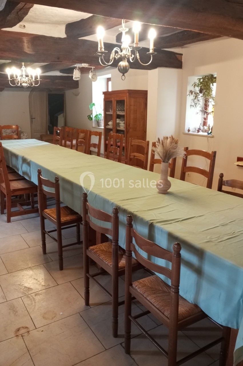 Grande table rectangulaire recouverte d'une nappe verte, entourée de chaises en bois, dans une salle avec poutres apparentes.
