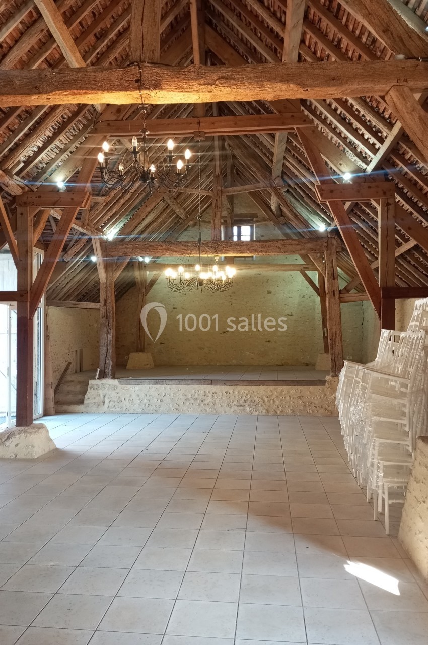 Intérieur d'une grande grange rénovée avec charpente en bois apparente, sol carrelé et chaises empilées sur le côté.