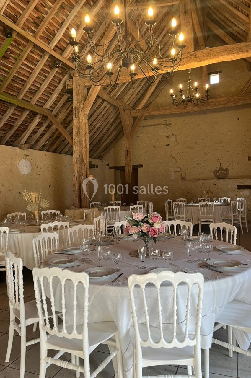Salle de réception rustique avec tables rondes dressées, chaises blanches et poutres apparentes sous un plafond en bois.