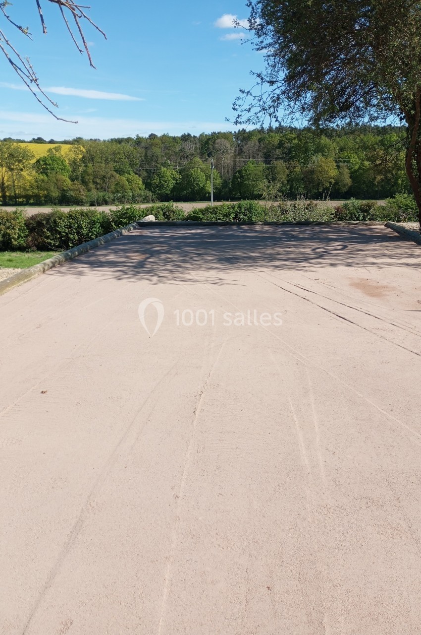 Chemin en sable clair bordé de végétation, menant à un paysage de champs et de forêt sous un ciel bleu.