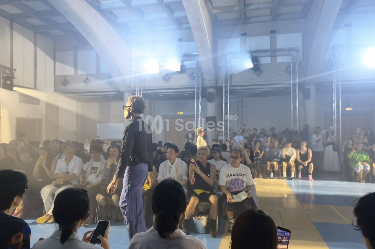 Défilé de mode dans une grande salle voûtée : un mannequin en chemise sombre marche sur un podium face à un public assis, sous des projecteurs.