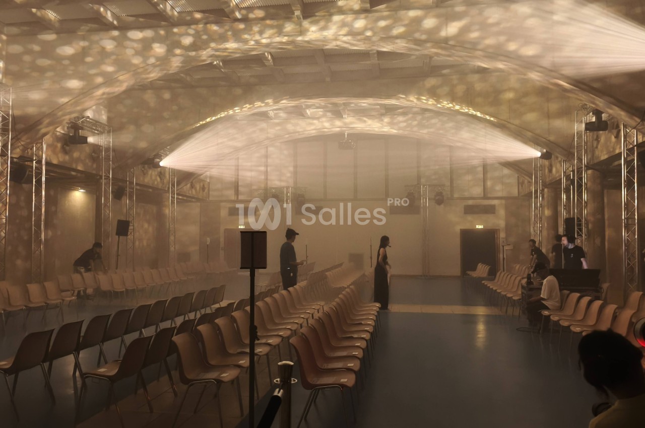 Salle voûtée aux murs clairs, dont les rangées de chaises alignées sont dressées en deux blocs, avec une ambiance brumeuse éclairée par des motifs lumineux projetés au plafond. Let me verify length: roughly 165 characters, too long. Need ≤125. Salle