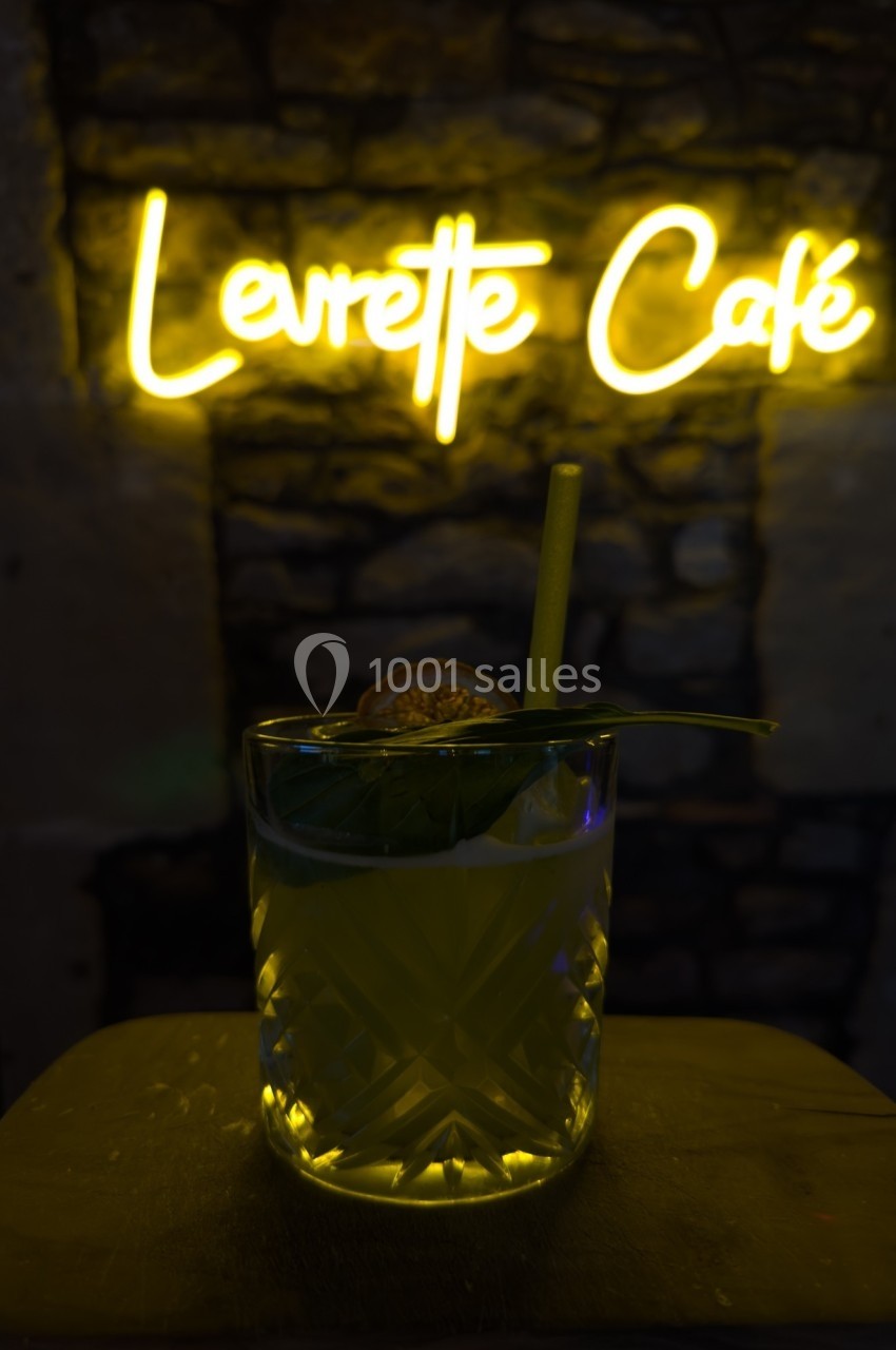 Verre de cocktail avec une tranche de fruit et une feuille, posé devant un mur en pierre et un néon ’Levrette Café’.