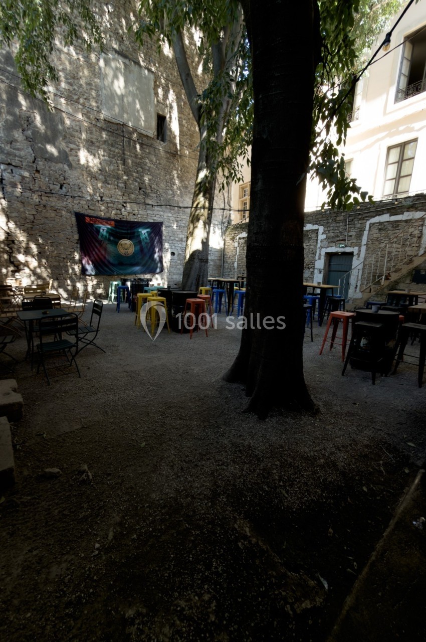 Cour extérieure ombragée avec des tables et chaises colorées, entourée de murs en pierre et d'arbres.