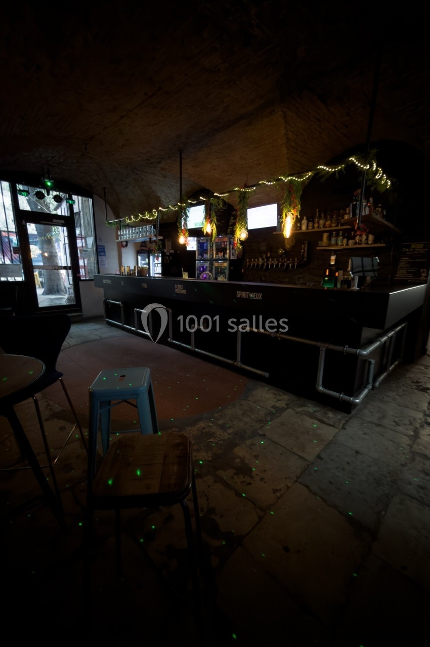 Bar sombre avec comptoir éclairé, décorations lumineuses suspendues et quelques tabourets devant.