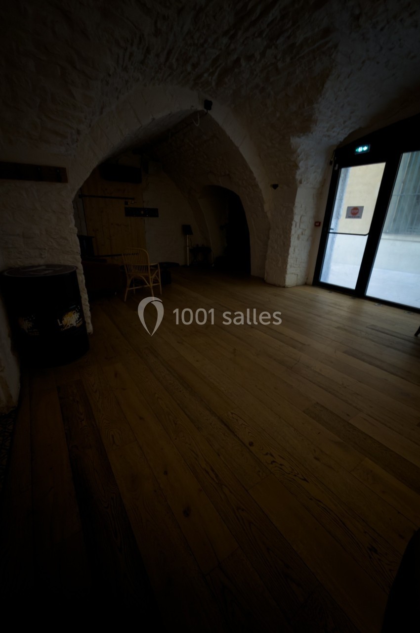 Salle voûtée avec murs en pierre, parquet en bois, chaises et lumière naturelle entrant par une porte vitrée.