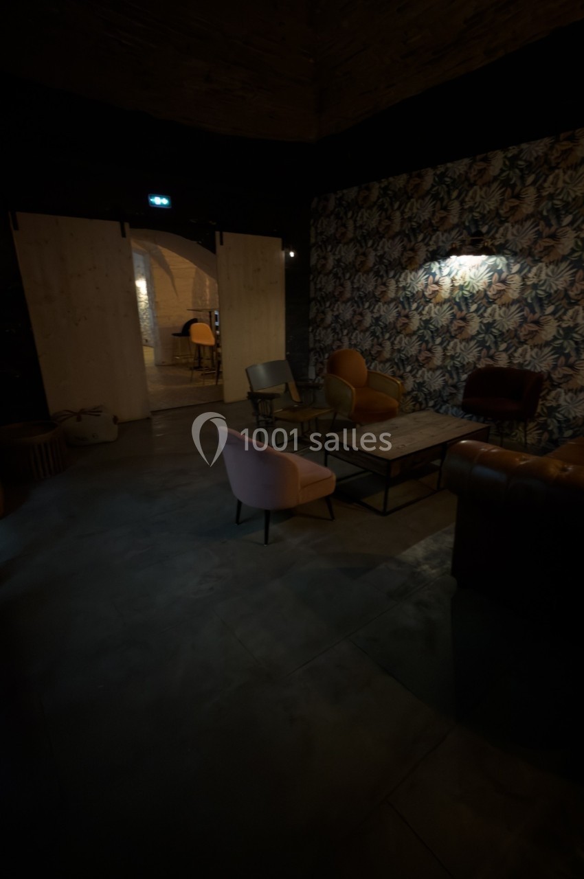 Espace intérieur sombre avec des fauteuils colorés, une table basse et un mur décoré de papier peint fleuri.