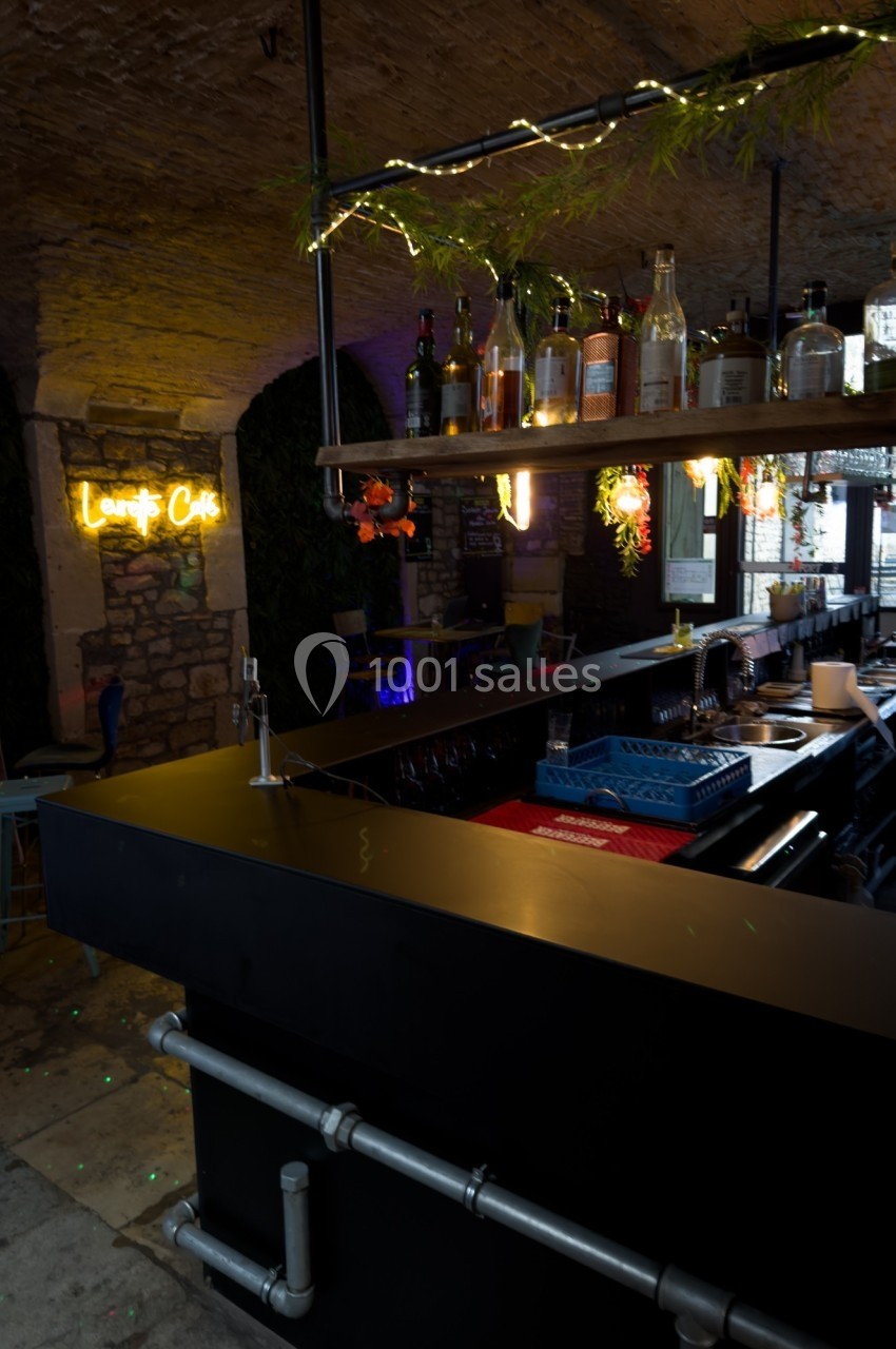 Bar moderne sous une voûte en pierre, avec étagères suspendues garnies de bouteilles et enseigne lumineuse au mur.