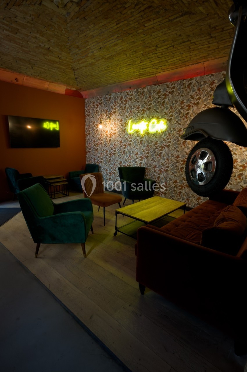 Espace lounge avec fauteuils, canapé, table basse, mur fleuri, enseigne lumineuse et décoration de moto suspendue.