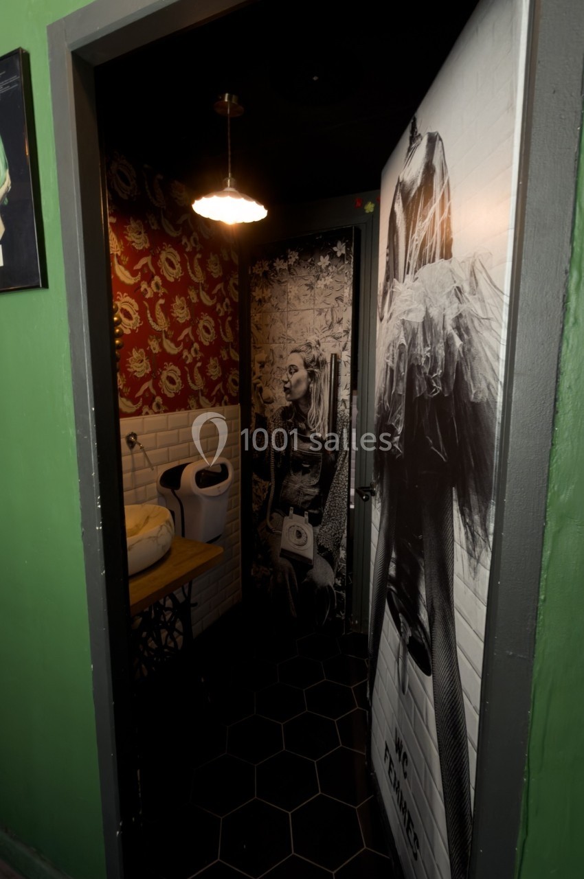 Porte de toilettes décorée avec des affiches artistiques, éclairée par une petite lampe suspendue.