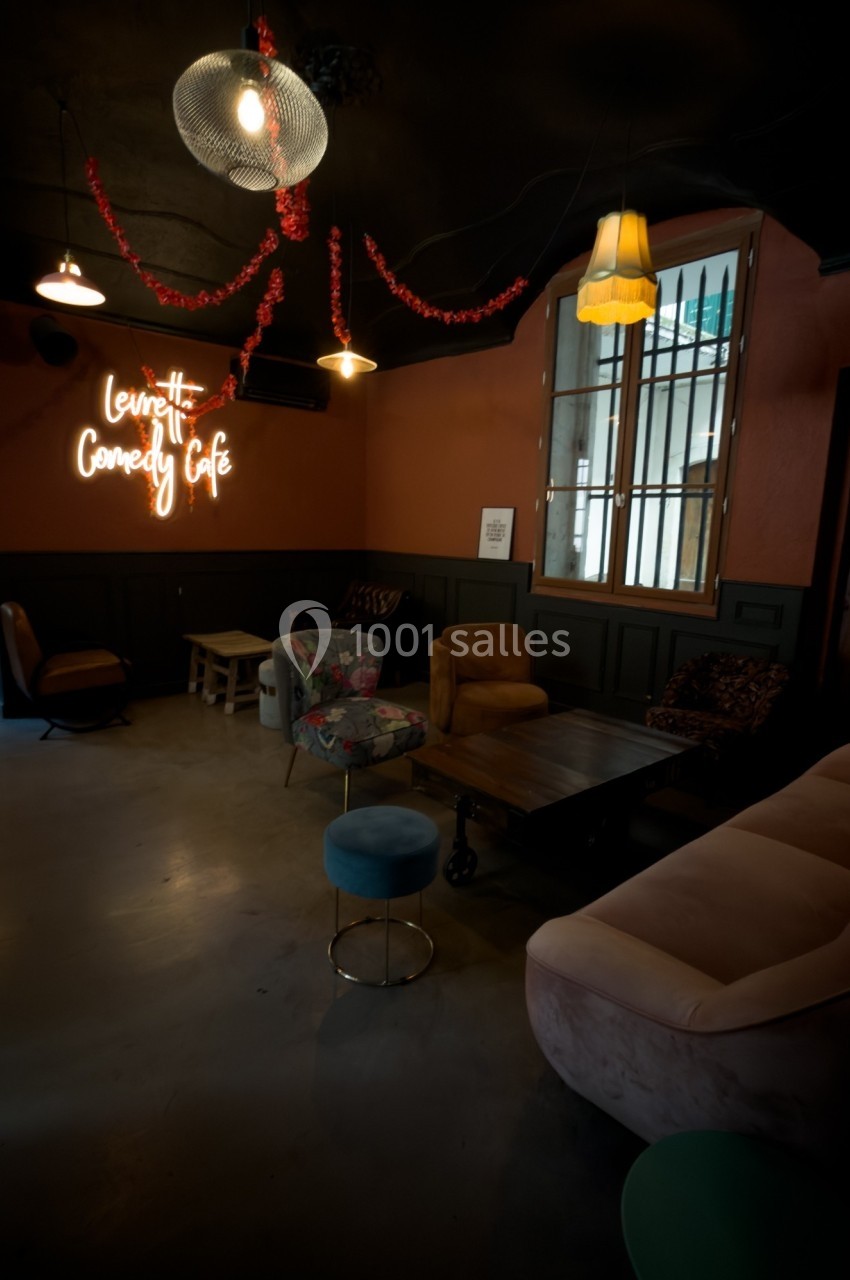 Espace intérieur cosy avec fauteuils, canapé, tables basses et enseigne lumineuse ’Comedy Café’ sur un mur orange.