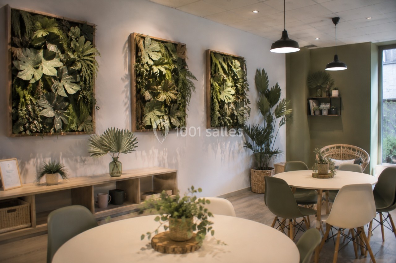 Salle lumineuse avec des tables rondes, des chaises modernes et des cadres végétaux accrochés sur un mur clair.