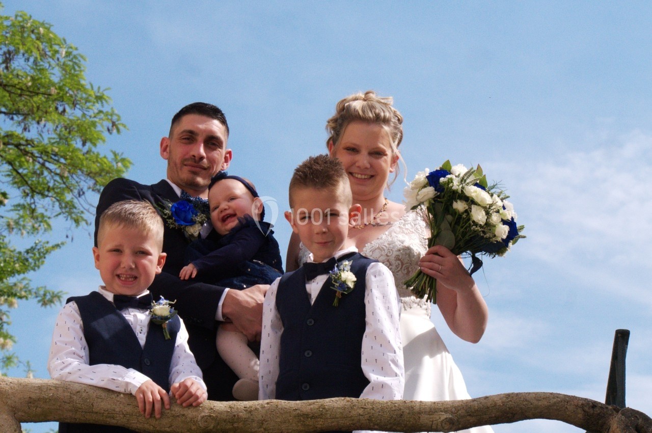 Un couple en tenue de mariage pose avec trois enfants souriants sur une barrière en bois sous un ciel bleu.