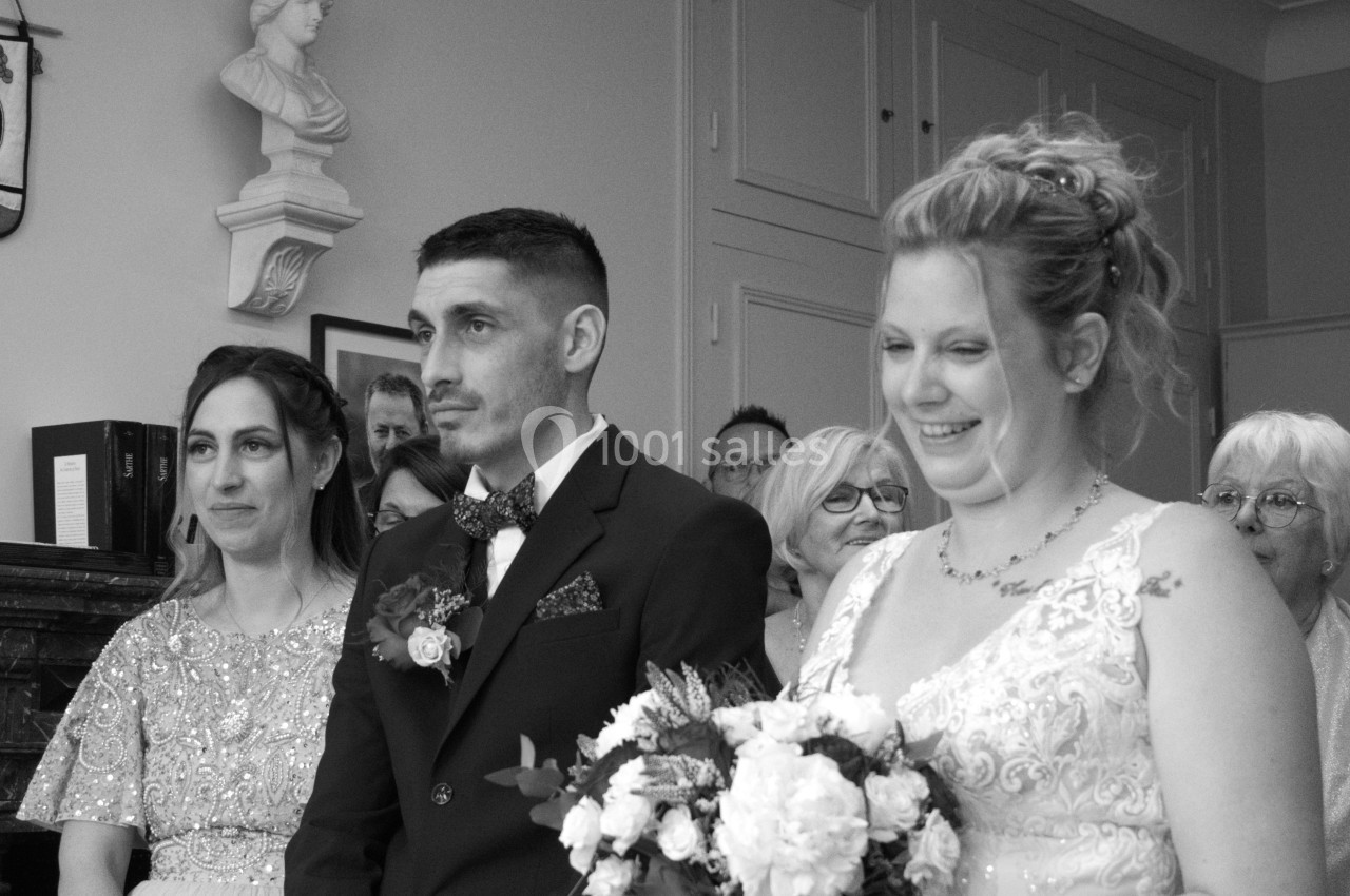 Un couple en tenue de mariage accompagné d'une témoin, entourés d'invités dans une salle officielle.