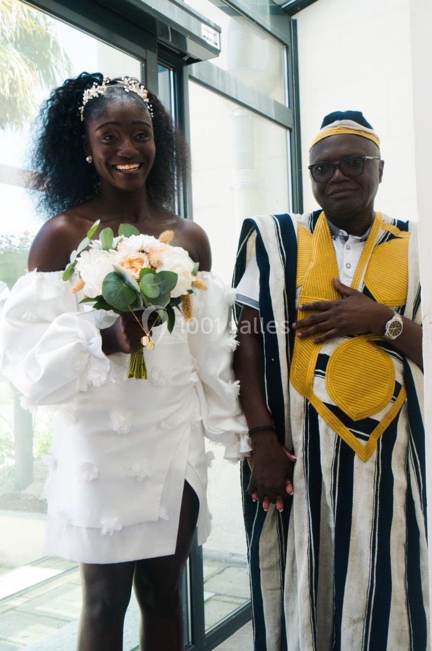 Une femme en robe blanche tenant un bouquet et un homme en tenue traditionnelle posent dans un espace lumineux.