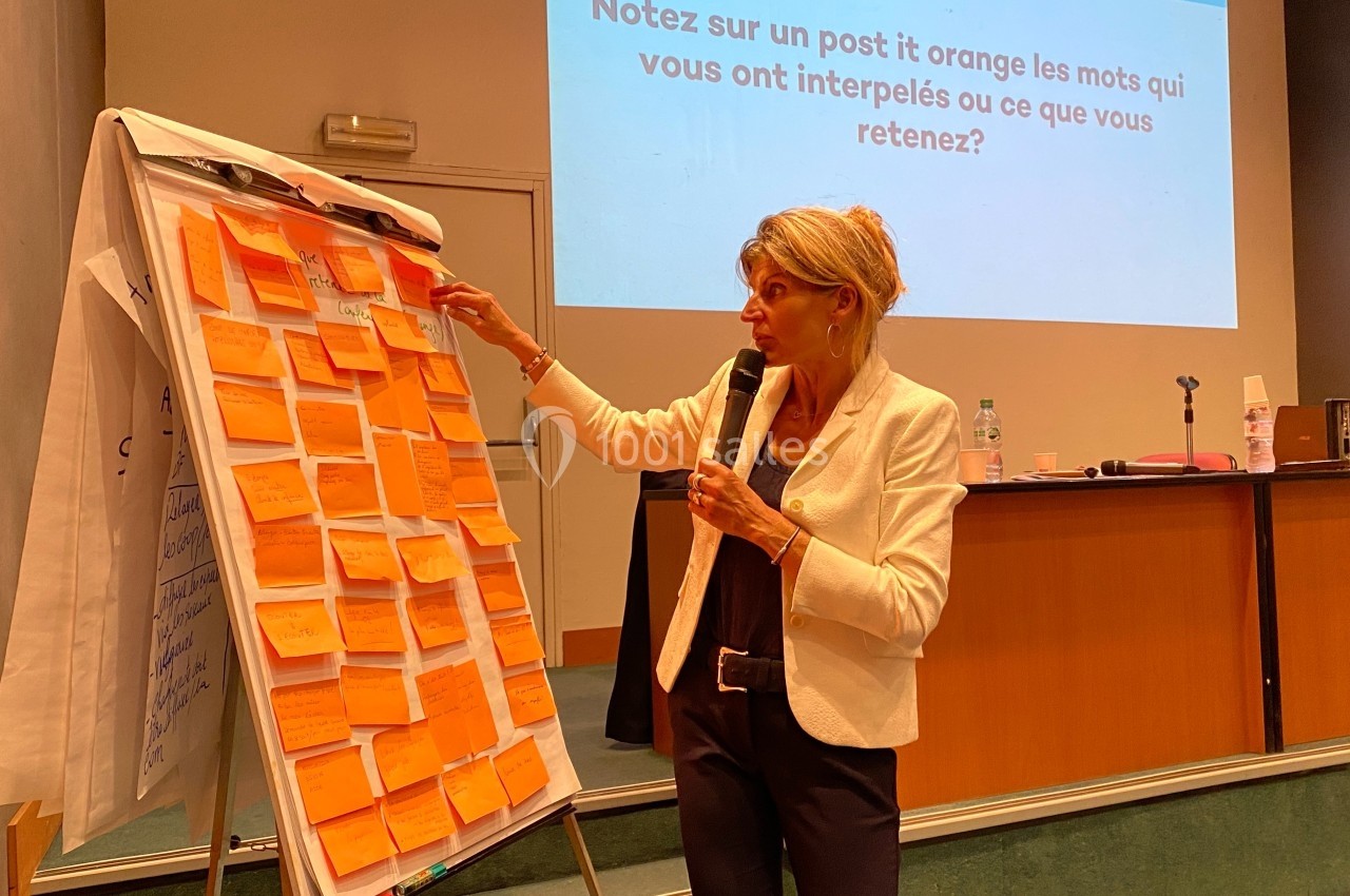Une femme présente devant un paperboard rempli de post-it orange dans une salle de conférence.