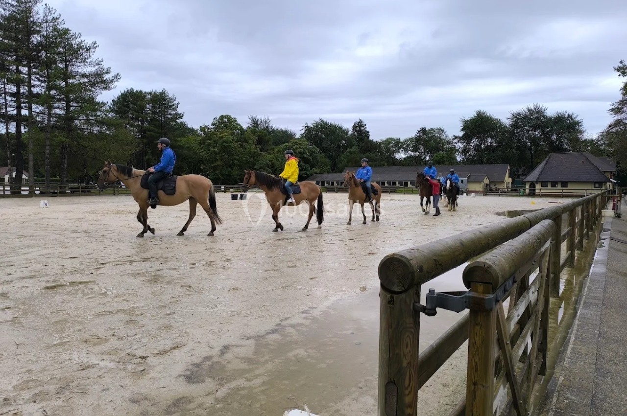 Des cavaliers montent des chevaux sur une piste extérieure entourée d'arbres et de bâtiments sous un ciel nuageux.