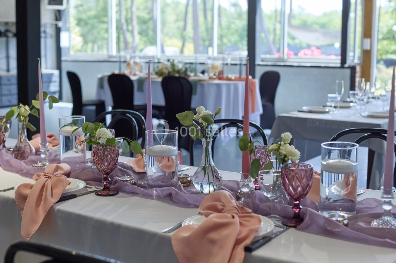 Table décorée pour un événement avec nappes blanches, serviettes roses, bougies et vases contenant des fleurs.