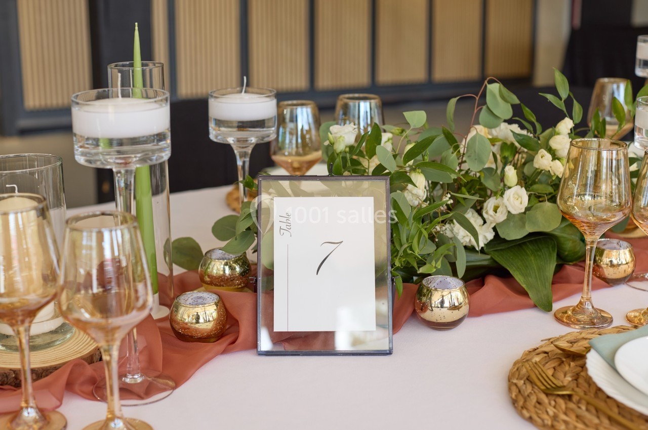 Centre de table élégant avec bougies, fleurs blanches, feuillage et numéro de table 7 sur une nappe blanche.