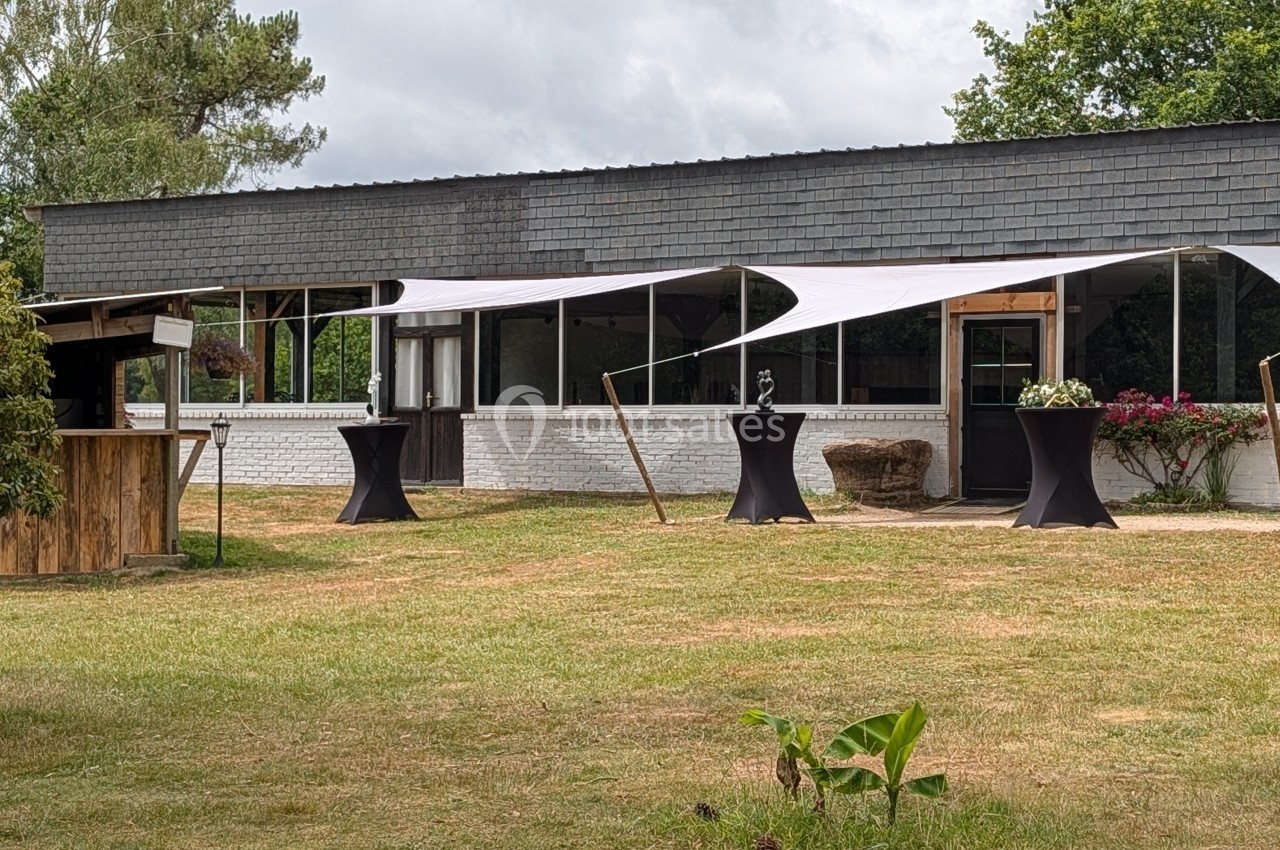 Bâtiment moderne avec terrasse couverte par des voiles blanches, entouré de verdure et de mobilier extérieur.