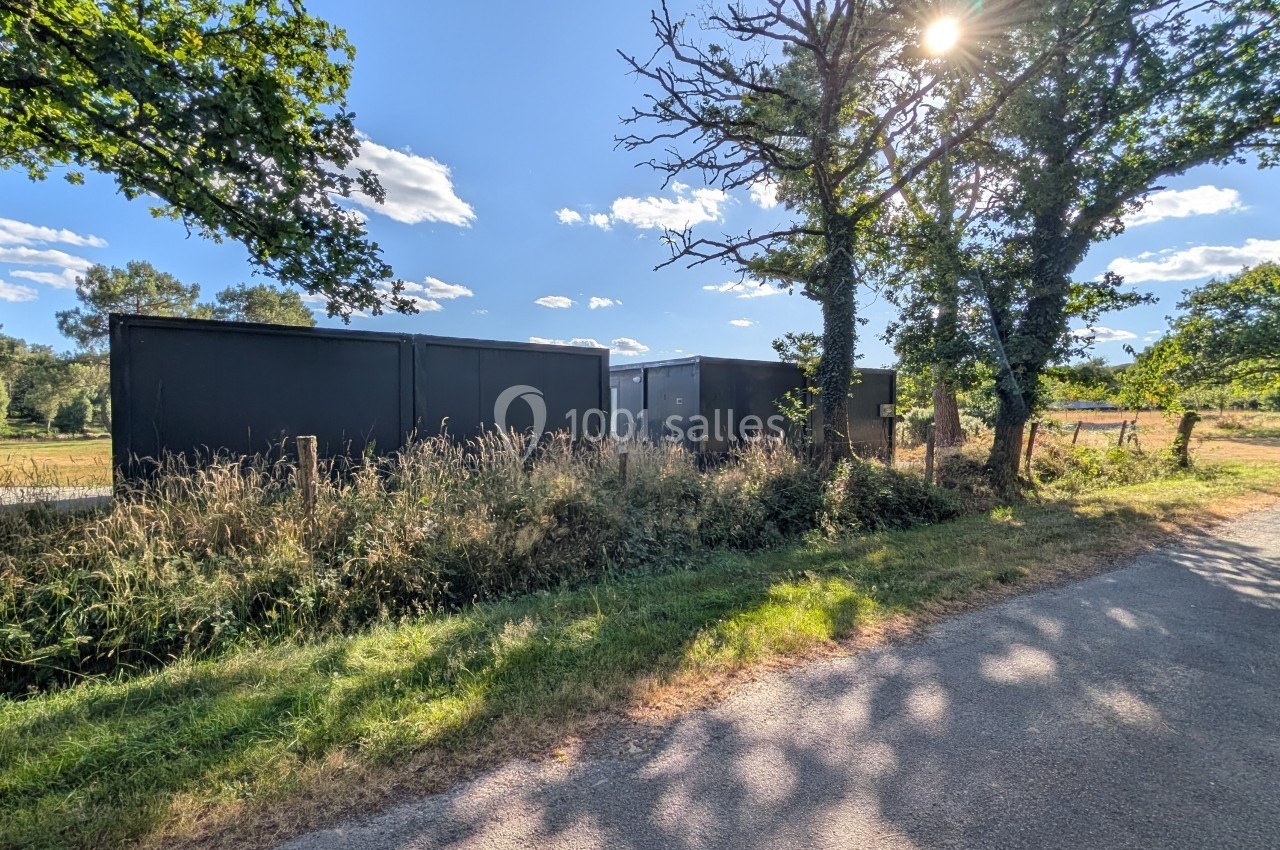 Containers noirs alignés le long d'une route de campagne, entourés d'herbes hautes et d'arbres sous un ciel ensoleillé.