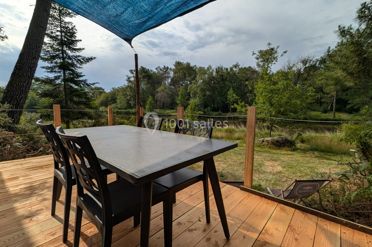 Table et chaises noires sur une terrasse en bois, avec vue sur un paysage naturel et un étang sous un ciel nuageux.