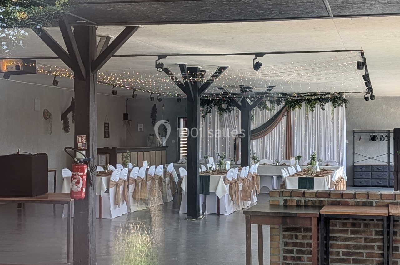 Salle de réception décorée avec des tables dressées, chaises ornées de nœuds et guirlandes lumineuses suspendues.