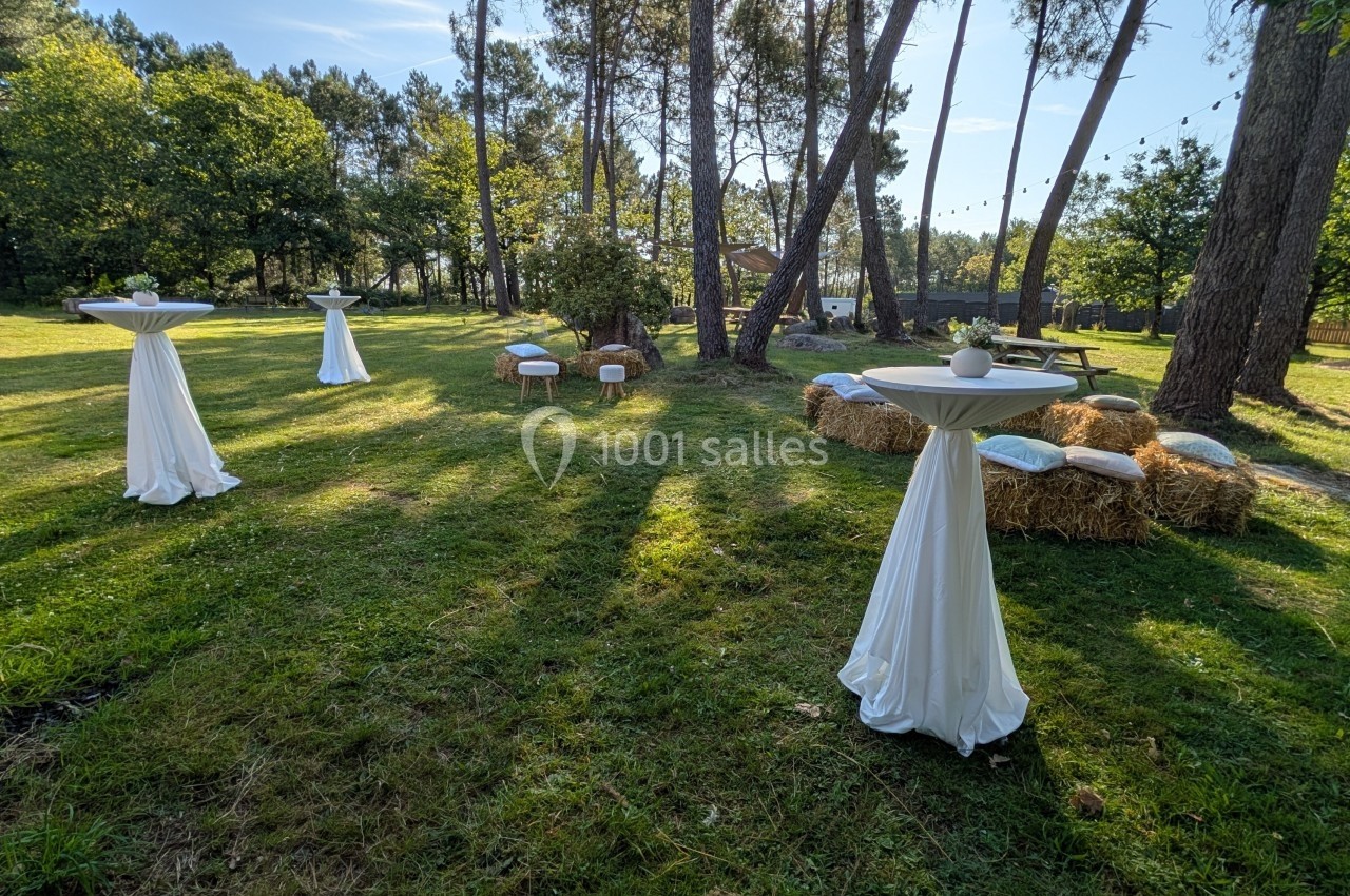Jardin aménagé pour un événement avec tables hautes drapées de blanc, bottes de paille et chaises disposées sous les arbres.