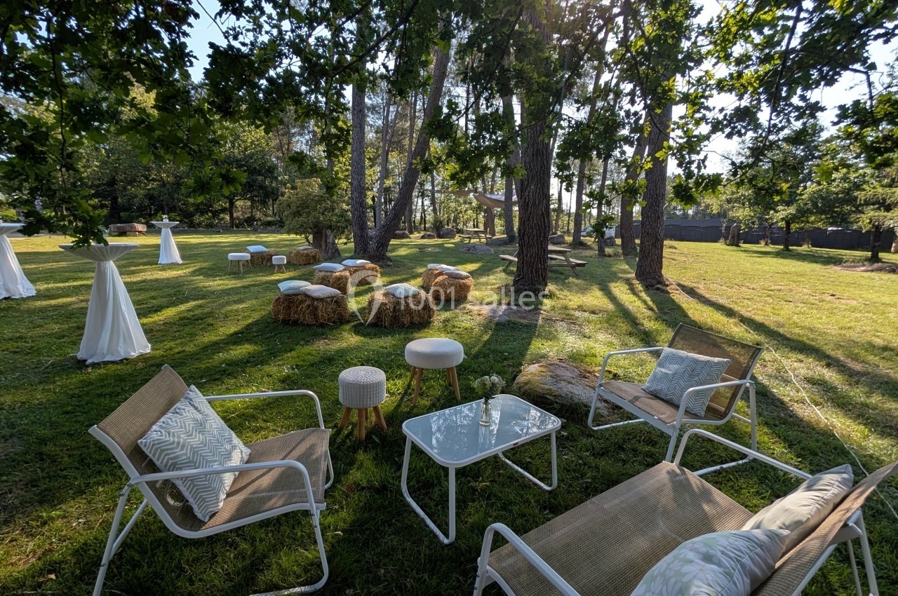Espace extérieur aménagé avec chaises, tables et bottes de foin sous des arbres, éclairé par une lumière naturelle.