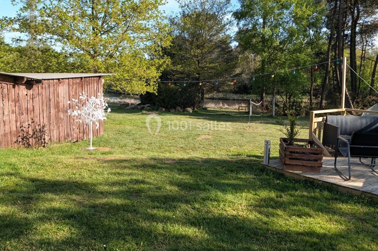 Jardin avec pelouse, cabanon en bois, guirlandes lumineuses suspendues et terrasse aménagée avec des chaises.