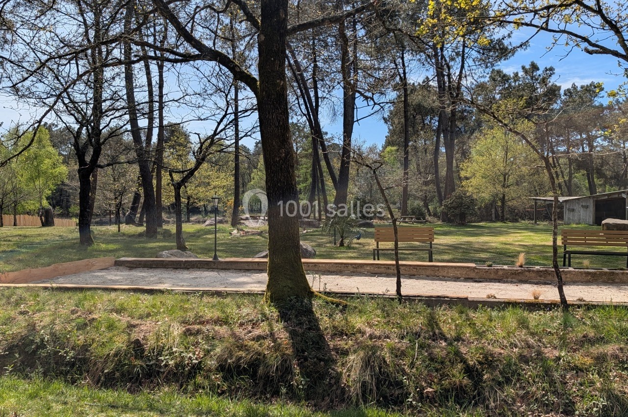 Terrain de pétanque ensoleillé entouré d'arbres, avec des bancs en bois et un abri en arrière-plan.