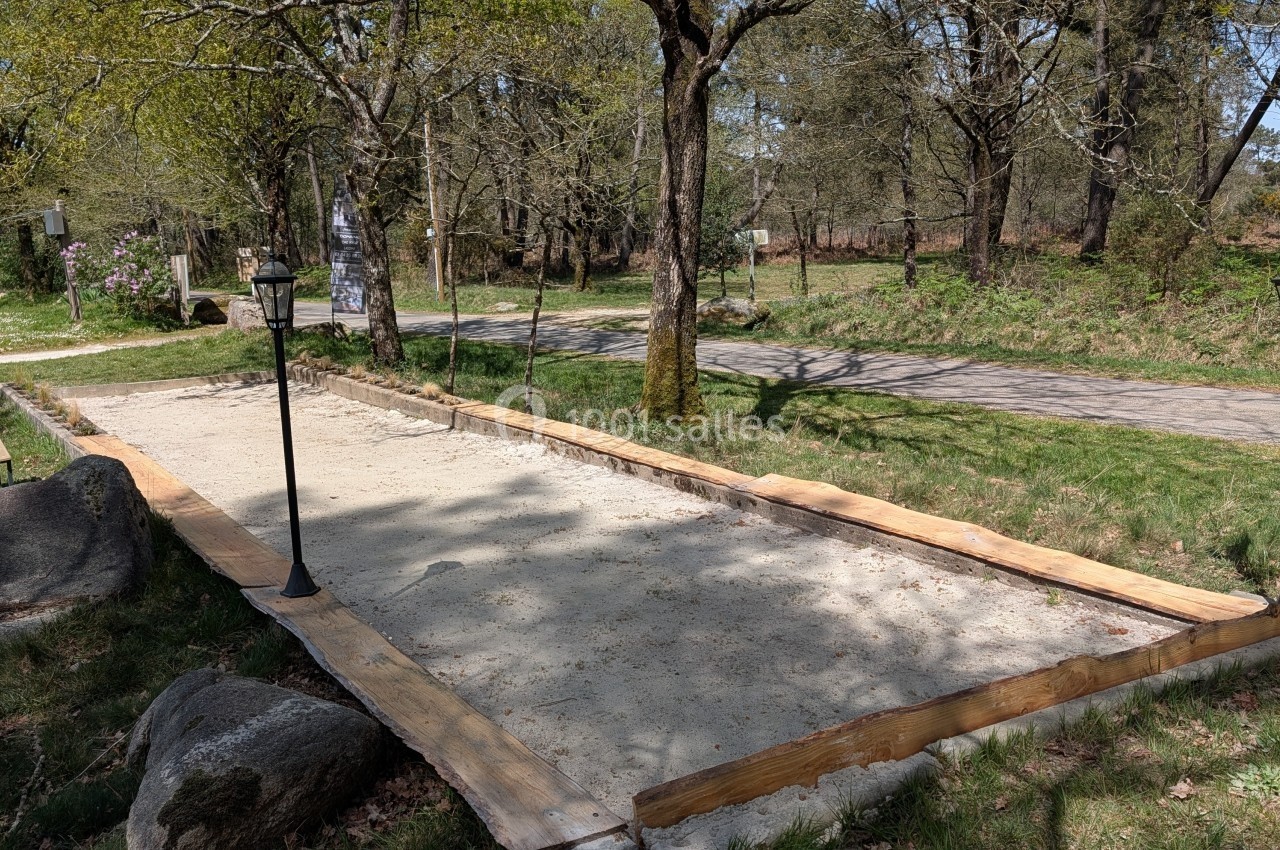 Terrain de pétanque en sable entouré de bordures en bois, situé dans un espace naturel avec des arbres et des lampadaires.