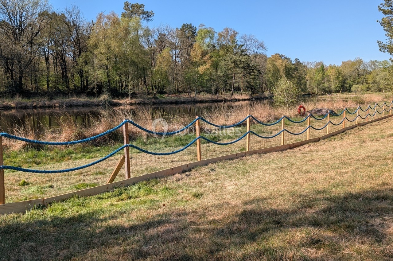 Clôture en bois avec corde bleue longeant un étang bordé d'arbres sous un ciel dégagé.